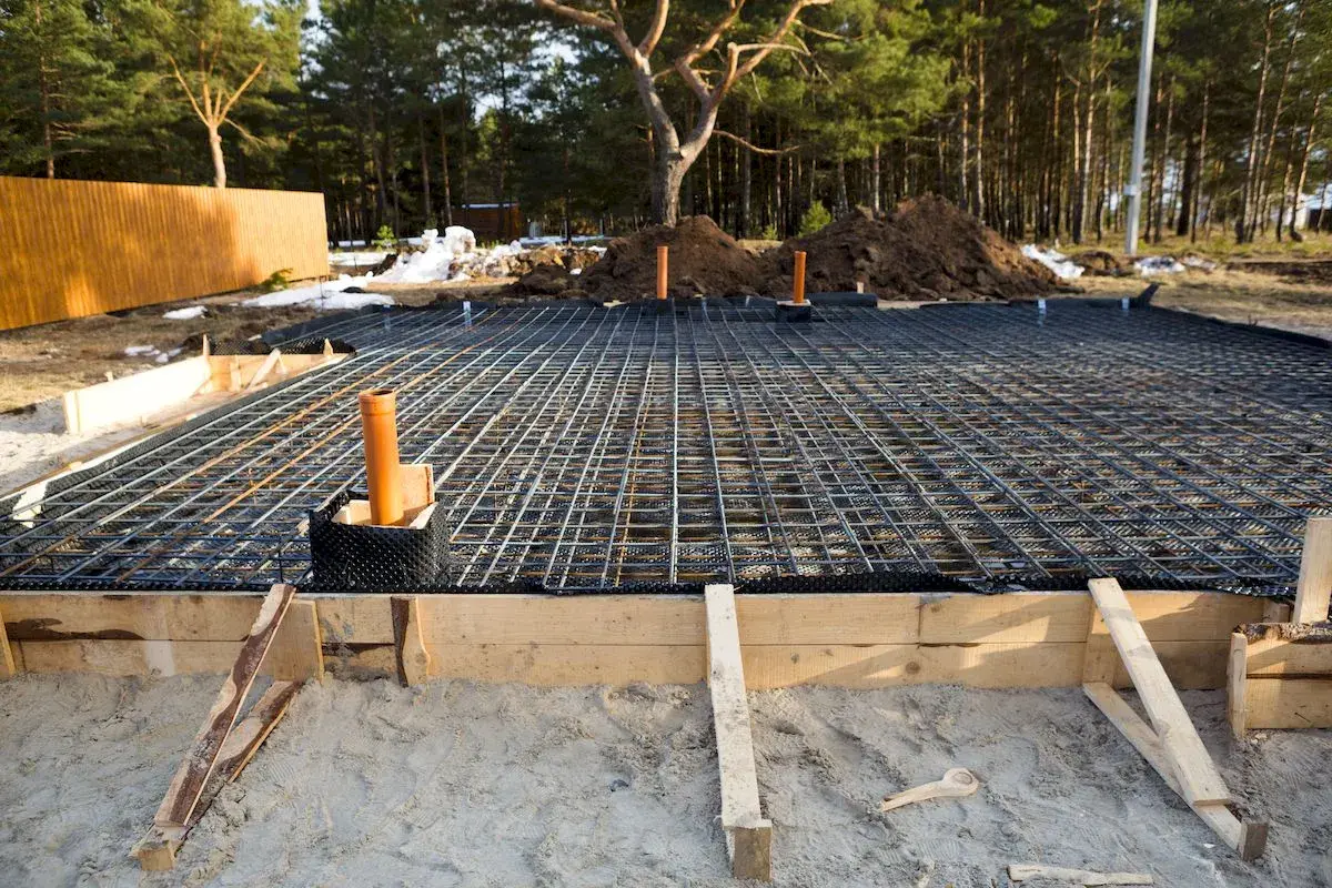 Gotowe zbrojenie płyty fundamentowej czeka na beton. Widać drewniane szalunki i pomarańczowe rury.