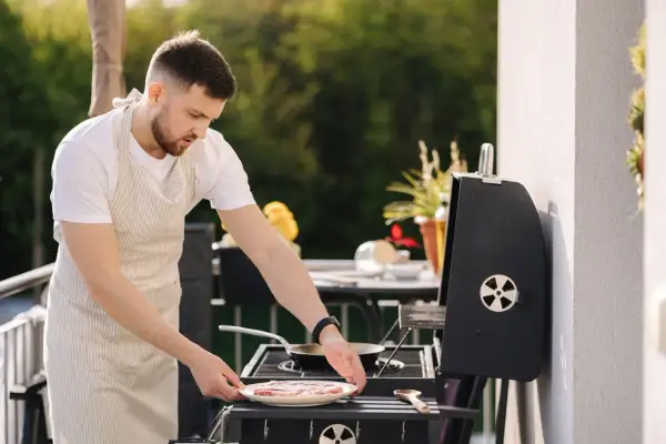 Mężczyzna w fartuchu przygotowuje mięso na grillu na balkonie. Czy można robić grilla na balkonie? Tak, jeśli jest to bezpieczne i zgodne z przepisami.